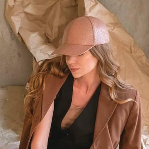 Może przedstawiać: Jasnobrązowa czapka z daszkiem o teksturowanej powierzchni. Czapka jest noszona przez osobę ubraną w brązową marynarkę na czarnym topie. Tło przedstawia pognieciony brązowy papier.