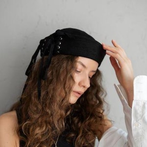 Op de afbeelding: Een zwarte hoofddoek met oogjes en strikjes. De hoofdbedekking is gemaakt van een geweven materiaal en wordt op het hoofd gedragen. De hoofddoek is een stijlvol accessoire.