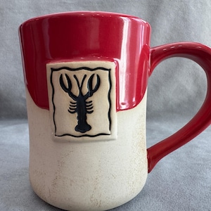 Puede incluir: Taza de cerámica con la parte superior y el asa rojas, y una base color crema. Un diseño de langosta negra aparece en un cuadrado color crema en la parte delantera de la taza. La taza está diseñada para bebidas calientes.