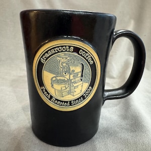 May include: Black coffee mug with a circular logo that reads "grassroots coffee" and "Fresh Roasted Since 2009". The logo features an illustration of a coffee roaster. The mug has a curved handle.