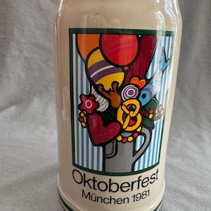 Puede incluir: Jarra de cerveza de cerámica color crema con un gráfico colorido de globos, pretzels y corazones. El diseño está sobre un fondo a rayas azules y blancas. La jarra está etiquetada como "Oktoberfest München 1981".