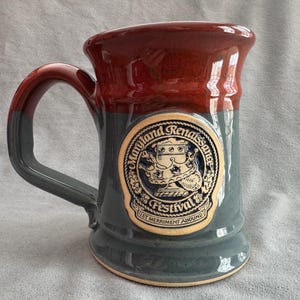 Può includere: Tazza in ceramica con bordo e manico rosso scuro, che sfuma in un corpo grigio. La tazza presenta un emblema del Maryland Renaissance Festival. L'emblema è un design circolare con testo e illustrazione decorativa.