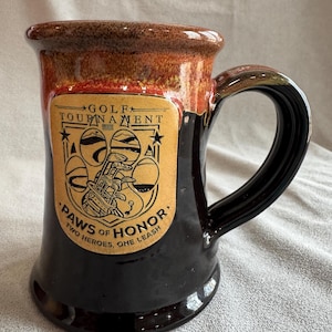 May include: A ceramic mug with a dark brown base and a lighter brown rim. The mug features a rectangular label with the words "Golf Tournament" and "Paws of Honor: Two Heroes, One Leash". The handle is black.