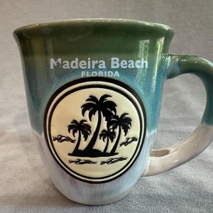 May include: A ceramic mug with a green and blue ombre glaze featuring the text Madeira Beach FLORIDA in white. A circular emblem on the front displays a black silhouette of palm trees against a cream background with small wave details.