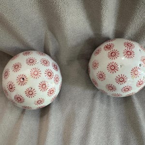 May include: Two spherical ceramic salt and pepper shakers featuring a white base with a repeating pattern of small red starburst or floral designs. Each shaker has a series of small holes at the top for dispensing seasonings.