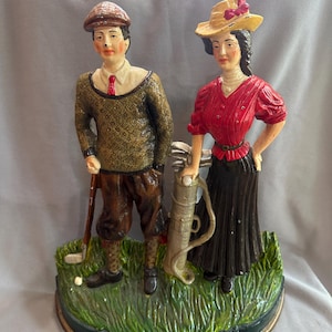 Puede incluir: Figura vintage de hierro fundido con temática de golf que representa a un hombre y una mujer con ropa de época. El hombre lleva gorra, jersey y pantalones bombachos, sosteniendo un palo de golf. La mujer viste blusa roja y falda negra, con bolsa de golf.