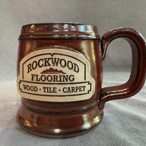 Può includere: Una tazza in ceramica marrone con finitura lucida e un grande manico. Una placca rettangolare color crema è centrata sulla parte anteriore, con il testo nero: ROCKWOOD FLOORING WOOD • TILE • CARPET. La tazza presenta una base larga e bordo spesso.