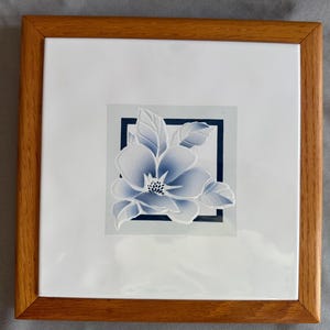 May include: Framed artwork with a blue and white floral design. The flower is within a dark blue square on a white background. The frame is made of light brown wood. The artwork is square in shape.