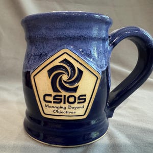 Puede incluir: Una taza de cerámica azul con un asa grande. La taza presenta un logotipo con el texto "CSIOS Managing Beyond Objectives" en un emblema color crema. El borde superior tiene un esmalte azul claro.
