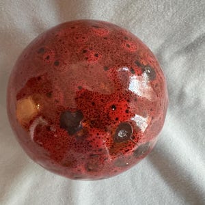 Könnte beinhalten: Runder, keramischer Dekorationsgegenstand mit glänzender Oberfläche. Die Oberfläche ist hauptsächlich rot mit dunkleren roten und braunen Flecken. Das Objekt hat eine glatte, kugelförmige Form.