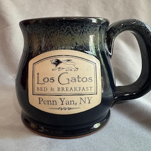 May include: A dark blue ceramic mug with a large handle. The mug features a cream-coloured label with the words "Los Gatos Bed & Breakfast" and "Penn Yan, NY" printed on it, along with a lion illustration. The mug has a glossy finish.
