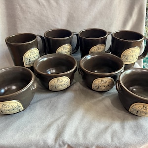 May include: A set of eight brown ceramic mugs and bowls. The mugs have handles and the bowls have small side handles. Each piece has a cream-colored label with the text "Billy Graham" and a landscape illustration.