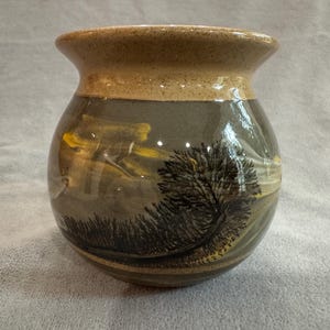 May include: A handcrafted ceramic vase with a landscape design. The vase features a rounded body and a slightly flared rim. The landscape design includes a tree, grass, and a yellow and gray sky. The vase is approximately 6 inches tall.