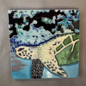May include: A square ceramic tile featuring a raised relief design of a sea turtle swimming. The turtle has a cream-colored head and flipper with dark spots, set against a background of glossy blue, green, and deep black abstract water patterns.