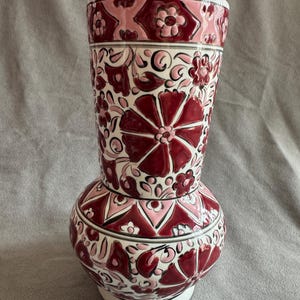Puede incluir: Un jarrón decorativo de cerámica con base bulbosa y parte superior cilíndrica. El jarrón presenta un estampado floral en tonos rojos, rosas y blancos. El diseño incluye flores estilizadas y formas geométricas. El jarrón tiene base y borde blancos.