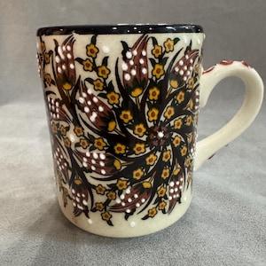 May include: A ceramic mug with a cream-colored base featuring a hand-painted floral pattern of dark red tulips and small yellow flowers. The design includes raised white dot accents, a dark rim, and a cream-colored handle with red dot details.