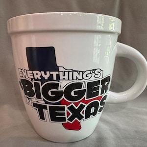 Puede incluir: Taza de cerámica blanca con un asa grande. La taza presenta la frase "Everything's Bigger in Texas" en letras negras y rojas, con un contorno azul y rojo de Texas. Un recuerdo o regalo divertido.