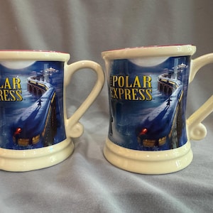May include: Two cream-colored ceramic mugs featuring a blue night scene of a train traveling across a bridge. The text POLAR EXPRESS is printed in yellow on the front. Each mug has a curved handle and a red interior rim, showcasing a winter landscape.
