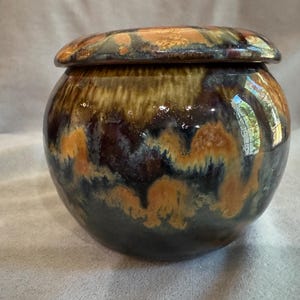 May include: A round ceramic jar with a matching lid, featuring a glossy glaze in shades of deep brown, black, and warm amber. The surface displays a fluid, dripped pattern where the colours blend together, creating a smooth, reflective finish.