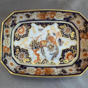 May include: A rectangular porcelain tray with rounded corners, featuring a white center with a bird and floral motif in orange, blue, and gold. The border is decorated with intricate floral patterns in deep blue, orange, and gold, finished with a gold-trimmed edge.