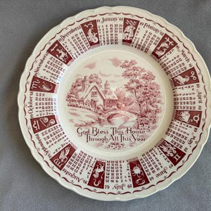 Könnte beinhalten: Ein weißer Teller mit gewelltem Rand und einem rot-weißen Design. Er zeigt einen Kalender für 1961, Tierkreiszeichen und ein zentrales Bild eines Hauses und einer Brücke. Der Text "God Bless This House Through All This Year" ist ebenfalls vorhanden.