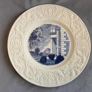 May include: A white decorative plate with a blue and white illustration of a building and trees. The plate has a detailed embossed border with floral and geometric designs. The central image is a detailed architectural scene.