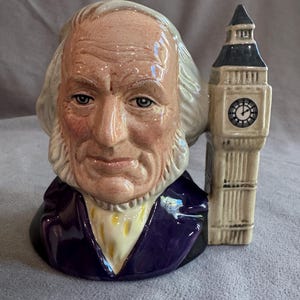 May include: A ceramic character jug shaped like an elderly man with white hair and sideburns, wearing a purple coat and yellow cravat. The handle of the mug is designed as a tall, beige clock tower with a dark roof and a circular clock face.