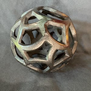 Puede incluir: Un objeto decorativo esférico con una estructura de celosía geométrica hueca. La superficie tiene un acabado metálico texturizado en tonos bronce y oro, con formas pentagonales y hexagonales interconectadas por toda la esfera.
