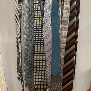 May include: A selection of neckties hanging on a white hanger. The ties feature diverse patterns, such as stripes, geometric shapes, and paisley designs. Colours include shades of brown, blue, grey, and teal.