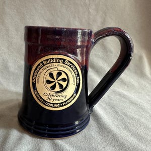 May include: A handcrafted ceramic mug with a dark blue exterior and a burgundy rim. The mug features a handle and a circular logo with the text "Advanced Building Services, Inc." and "Celebrating 20 years."