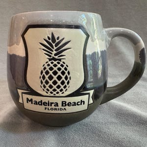 May include: A ceramic mug with a rounded shape featuring a cream-coloured shield emblem with a black pineapple graphic. Below the pineapple, a banner displays the text Madeira Beach FLORIDA in black lettering. The mug has a grey and blue glaze.