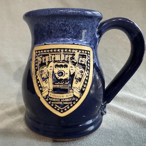 Puede incluir: Una taza de cerámica azul oscuro con un asa grande. La taza presenta un emblema dorado y negro con el texto "September Fest" y un diseño de tren. La taza es una pieza única de cristalería.