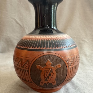 May include: A terracotta-colored vase with a glossy black neck and rim. The body features a central circular medallion with a carved turtle motif, surrounded by textured geometric patterns and horizontal bands in black and natural clay tones.