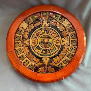 May include: A circular wooden wall plaque featuring a detailed, multi-layered relief carving of the Aztec sun stone calendar. The design includes a central face with a protruding tongue, surrounded by intricate geometric patterns in shades of brown.
