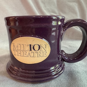 Puede incluir: Una taza de cerámica de color púrpura oscuro con un asa grande. La taza presenta las palabras "MILTON THEATRE" en letras doradas sobre un óvalo color crema. La taza tiene un acabado brillante.