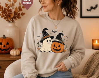 May include: A beige crewneck sweatshirt featuring a graphic print of a ghost wearing a witch hat standing next to a jack-o'-lantern wearing a witch hat, surrounded by small bats, stars, and autumn leaves, paired with light blue denim jeans.