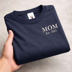 May include: Navy blue sweatshirt with the word "MOM" embroidered in a light beige color, along with "Est. 2023". The sweatshirt is folded and held by a hand, showcasing the design on the front. The background is a neutral gray.
