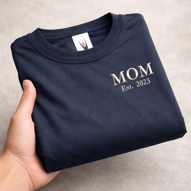 May include: Navy blue sweatshirt with the word "MOM" embroidered in a light beige color, along with "Est. 2023". The sweatshirt is folded and held by a hand, showcasing the design on the front. The background is a neutral gray.