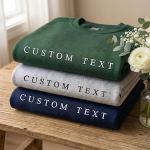 May include: Three folded sweatshirts in forest green, grey, and navy blue, each with the text "CUSTOM TEXT" in white. The sweatshirts are stacked on a wooden surface, with a vase of white flowers in the background.