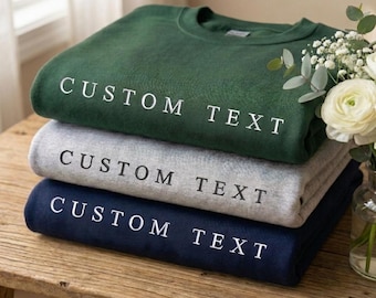 Könnte beinhalten: Drei gefaltete Sweatshirts in Tannengrün, Hellgrau und Marineblau, jeweils mit dem Text "CUSTOM TEXT" in Weiß. Die Sweatshirts sind auf einer Holzoberfläche gestapelt, mit einer Vase weißer Blumen im Hintergrund.