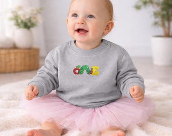 Könnte beinhalten: Ein graues Langarm-Sweatshirt mit einer Stickerei des Wortes ONE, bei dem der Buchstabe O ein roter Apfel, der Buchstabe N eine grüne Raupe und der Buchstabe E eine gelbe Form ist, kombiniert mit einem hellrosa Tüll-Tutu-Rock.