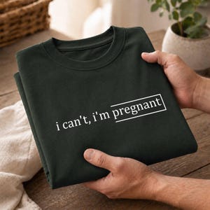 May include: A dark forest green crewneck sweatshirt featuring the white text "i can't, i'm pregnant" printed on the front. The word "pregnant" is enclosed within a white rectangular outline, creating a clean and simple graphic design on the fabric.