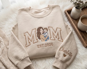 Könnte beinhalten: Cremefarbenes Sweatshirt mit gesticktem Design. Das Design zeigt das Wort "MOM" im College-Stil, mit einer Illustration einer Mutter und eines Kindes. Das Jahr "EST. 2026" befindet sich unter der Illustration, und der Name "Charlotte" ist auf dem Ärmel gestickt.
