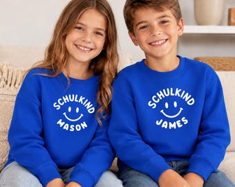 Könnte beinhalten: Zwei passende königsblaue Sweatshirts mit Rundhalsausschnitt und weißem Grafikdruck. Das Design zeigt den kreisförmigen Schriftzug SCHULKIND über einem Smiley, wobei die Namen MASON und JAMES jeweils unter dem Smiley aufgedruckt sind.