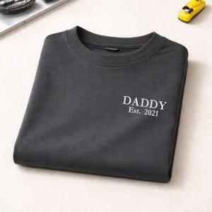 May include: Dark gray crew neck t-shirt with the word "DADDY" and "Est. 2021" printed in white. The shirt is folded neatly. A small yellow toy car is in the background.