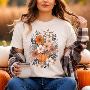以下が含まれることがあります： クリーム色のTシャツで、中央にカボチャと、オレンジ、ピーチ、ブラウン、落ち着いたグリーンの色合いの花や葉のブーケが描かれたグラフィックプリントがあり、丸首で半袖のデザインです。