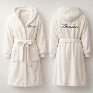 May include: Cream-coloured hooded robe with the name "Harrison" embroidered on the front and back. The robe features a tie belt, pockets, and a soft, plush texture. The robe is displayed from the front and back.