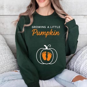 May include: A dark green long-sleeve sweatshirt featuring the white text "GROWING A LITTLE" above the orange text "Pumpkin". Below the text is a white outline of a pumpkin containing two orange baby footprints centred within the design.