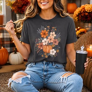 以下が含まれることがあります： チャコールグレーの半袖Tシャツ。中央にはカボチャと、その周りを囲むオレンジ、ピーチ、白の花々、そして緑の葉が描かれたグラフィックプリントが施されています。Tシャツは、ダメージ加工が施されたライトブルーのデニムジーンズと組み合わされています。
