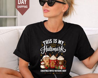 Könnte beinhalten: Ein schwarzes Kurzarm-T-Shirt mit einem Grafikdruck und dem Text "THIS IS MY Hallmark CHRISTMAS MOVIE WATCHING SHIRT". Das Design zeigt drei Tassen mit Heißgetränken, einen kleinen Schneemann sowie in rot, grün und weiß verpackte Geschenke.